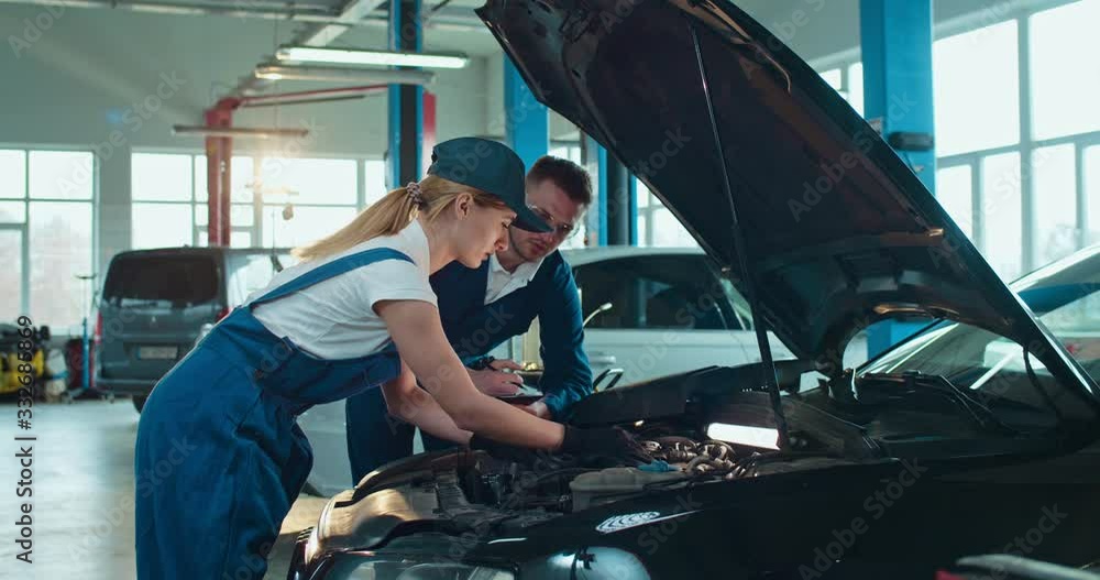 Caucasian man teaching young woman in overalls and hat to repair car in auto service salon. Female worker learning and repairing motor in big garage while male colleague helping and controlling. Caucasian man teaching young woman in overalls and hat to repair car in auto service salon. Female worker learning and repairing motor in big garage while male colleague helping and controlling.