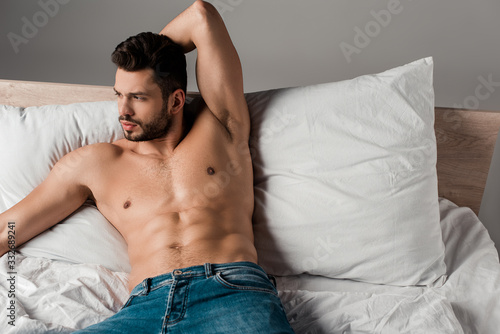brunette sexy shirtless man in jeans lying on bed on grey