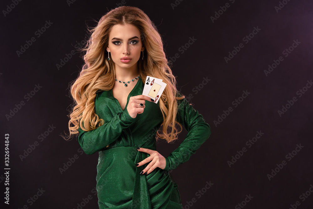 Blonde female in green stylish dress and jewelry. Put hand on hip, showing two playing cards, posing on black background. Poker, casino. Close-up Blonde female in green stylish dress and jewelry. Put hand on hip, showing two playing cards, posing on black background. Poker, casino. Close-up