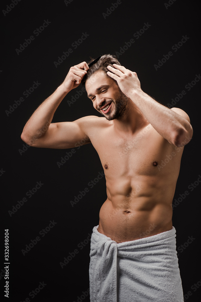 Fototapeta premium cheerful handsome man in towel combing hair isolated on black