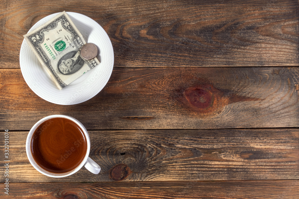 White cup with invigorating espresso coffee and saucer with two American dollar bill and coin. On dark wooden background. Payment or tip. Top view. Copy space for text. White cup with invigorating espresso coffee and saucer with two American dollar bill and coin. On dark wooden background. Payment or tip. Top view. Copy space for text.