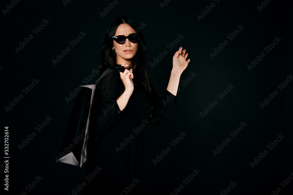 Beautiful picky girl in sunglasses and black dress with a shopping bag on her back makes funny face. Isolated on black Beautiful picky girl in sunglasses and black dress with a shopping bag on her back makes funny face. Isolated on black