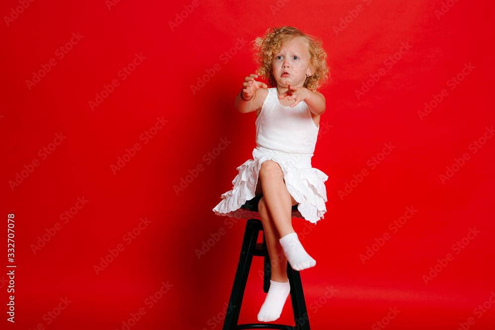 Amuzing funny little blonde girl in white dress sitting on chair wants something isolated on red Amuzing funny little blonde girl in white dress sitting on chair wants something isolated on red