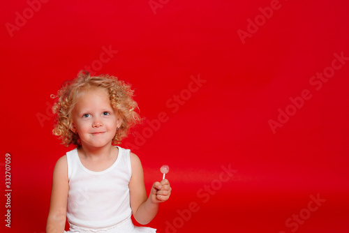 Beautiful little curly blonde girl eating lollipop sitting on the floor isolated on red