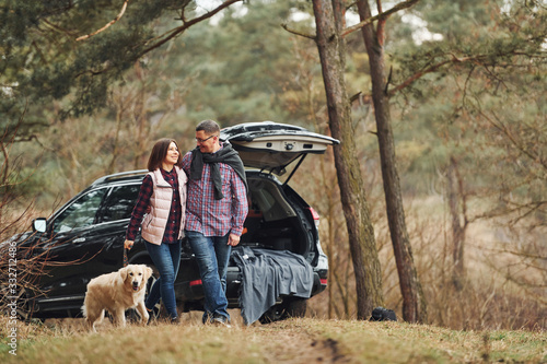 Happy mature couple have a walk with their dog in autumn or spring forest near modern car
