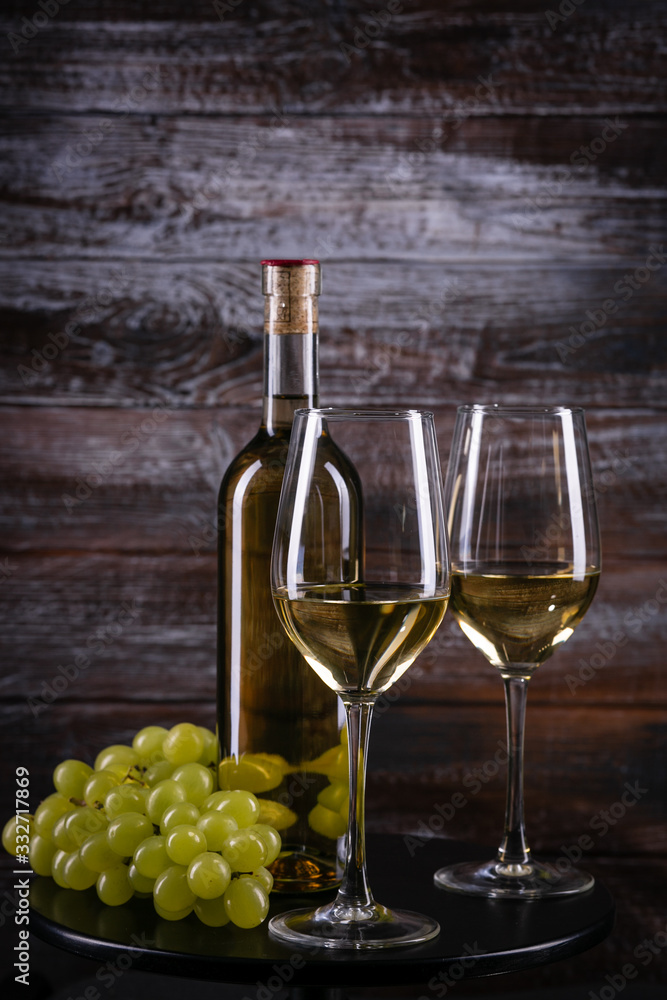White wine bottle and two glasses with grapes on a table on wooden background