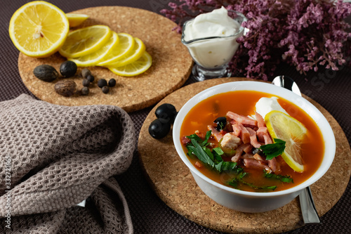 Soup Solyanka is a combined meat soup with smoked meat , various types of meat and tomato paste, serving dishes with sour cream, lemon and fresh herbs. Black olives on the table and a sprig of fresh f