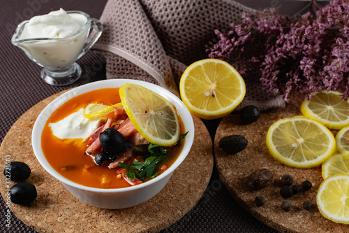 Soup Solyanka is a combined meat soup with smoked meat , various types of meat and tomato paste, serving dishes with sour cream, lemon and fresh herbs. Black olives on the table and a sprig of fresh f