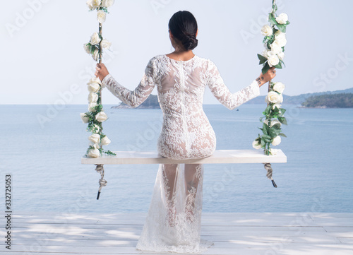 Back view of beautiful woman in white lace dress posing on the outdoor swing with flowwers over sea and sky background photo