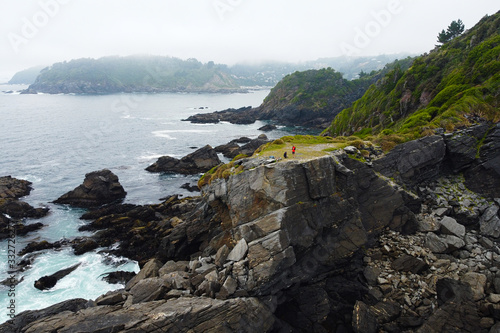 Stunning aerial view of travellers on staying on steep cliff. Drone view from above of jagged rocky coast of Pacific Ocean and rainy weather with fog. Bahia Mansa, Maicolpue, Osorno, Chile. photo