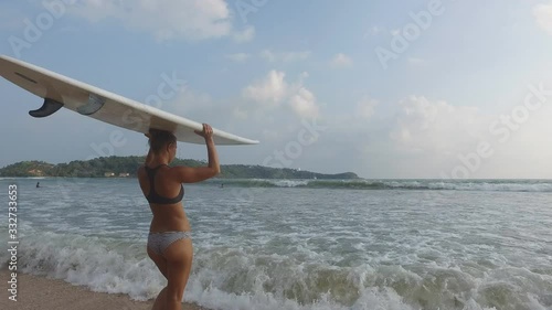 A young sexy surfer girl with a beautiful booty enters the ocean and carries a surfboard over her head. Live camera.