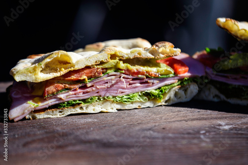 Fresh healthy sandwich or burger with ham, green salad and pickled cucumbers on a brown wooden table in direct sunlight at a street food market, side view photograph with soft focus 