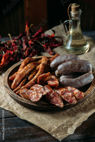 Traditional three kinds of meat sausages in rustic wood bowl, dried chili peppers and oil in a glass jug on natural black wood table.