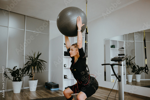 Slim woman in EMS training suit doing squats with a grey fitness ball, holding it above her head, side view.