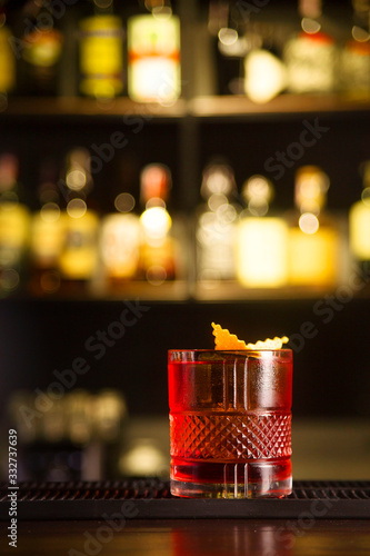 Valokuvatapetti Red cocktail with ice decorated with orange peel on a bar counter against the background of alcohol bottles
