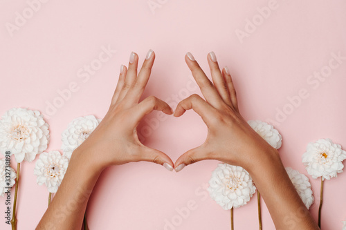 Woman's hands making a heart sign, among dahlia flowers on pink background. The concept of tenderness.