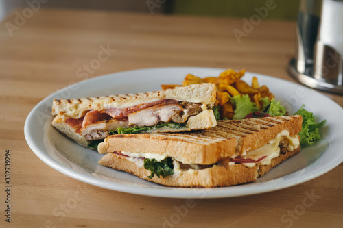 Close-up of chicken and bacon club sandwich with fries and vegetables on a white plate on a wooden background.