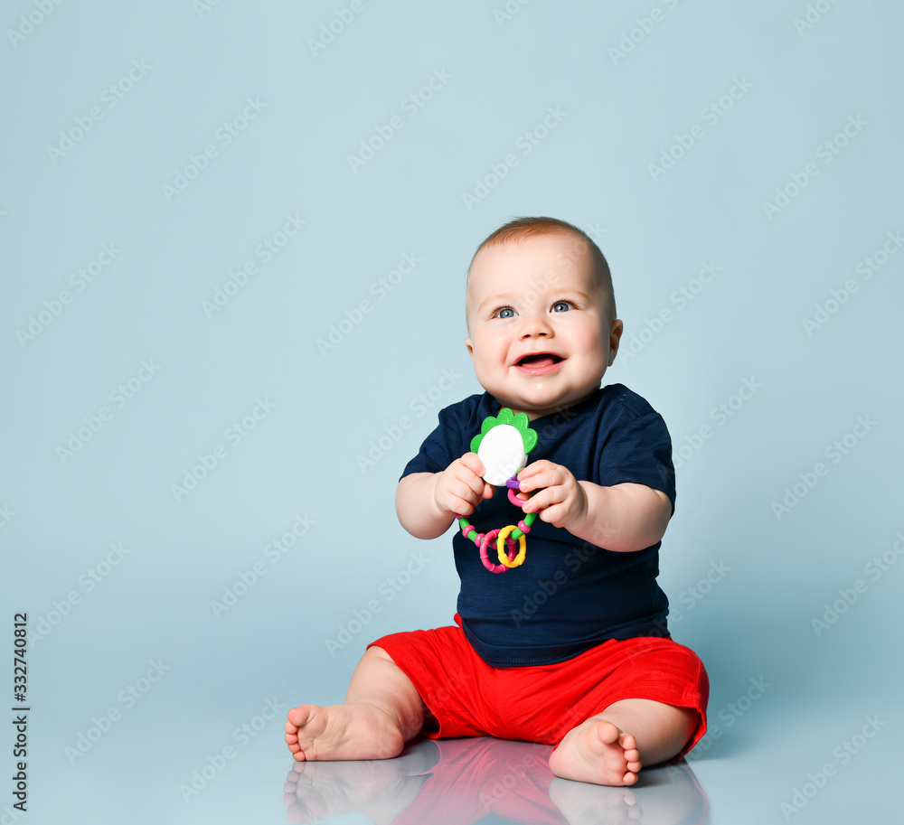 Toddler in t-shirt and red shorts, barefoot. He smiling, holding a rattle, sitting on floor against blue background. Close up Toddler in t-shirt and red shorts, barefoot. He smiling, holding a rattle, sitting on floor against blue background. Close up