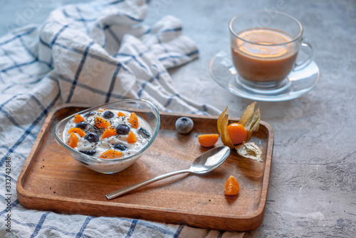 Breakfast with coffee and yogurt in a glass bowl with blueberries and physalis on a wooden brown tray on a concrete gray-blue background with a towel in a thin blue check.