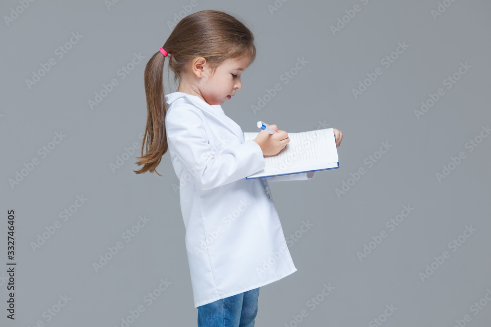 Adorable child girl uniformed as doctor is writing medical record isolated on grey background Adorable child girl uniformed as doctor is writing medical record isolated on grey background