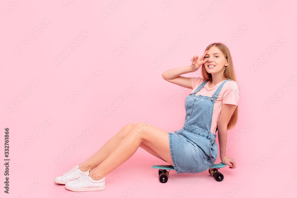 stylish attractive young woman sitting on a skateboard showing a peace gesture, on an isolated pink background stylish attractive young woman sitting on a skateboard showing a peace gesture, on an isolated pink background