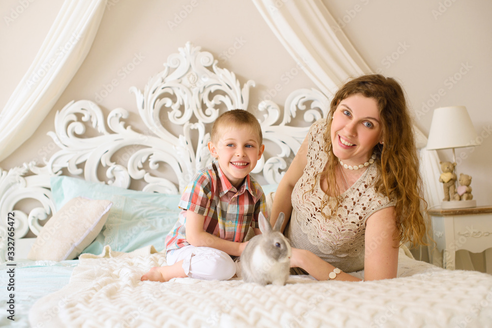 Happy mom with her son in light bedroom on bed playing with gray rabbit Happy mom with her son in light bedroom on bed playing with gray rabbit