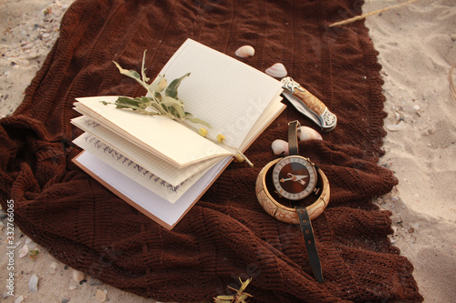 Notepad, compass, knife, olive branch, spikelet, bone bracelet and shells on a brown bedspread in the sand.