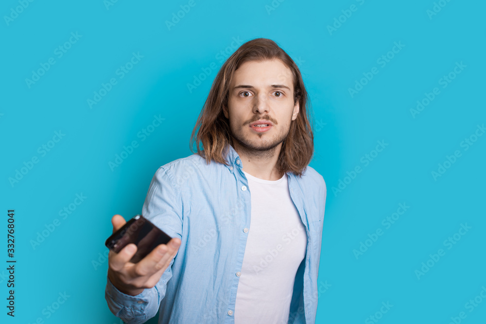 Long haired caucasian man with bristle is looking seriously at camera while holding a mobile on a blue background Long haired caucasian man with bristle is looking seriously at camera while holding a mobile on a blue background