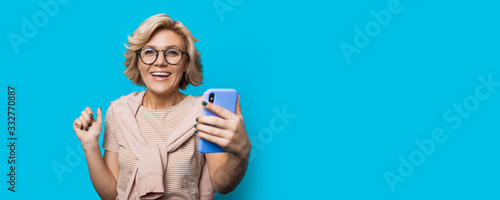 Blonde caucasian woman with eyeglasses is holding a phone and looking to the camera while posing on a blue wall with free space