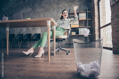 Full size photo of positive cheerful worker marketer girl have no work feel fun throw crumpled paper in trash bin sit chair in modern loft office