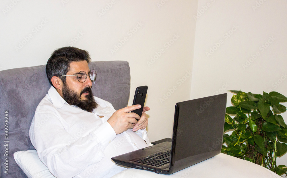 handsome bearded man wearing formal white shirt and sitting on bed with notebook in modern room. Distance work concept. Man surfing internet or working online. handsome bearded man wearing formal white shirt and sitting on bed with notebook in modern room. Distance work concept. Man surfing internet or working online.