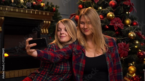 Two pretty young girls or women are sitting near a New Year or Christmas tree and taking selfie on a mobile phone.