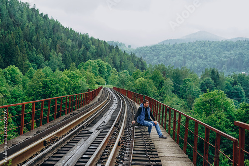 A man sits on a railway track in a blue jacket. Track on mountains background.