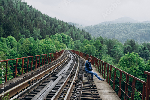 A man sits on a railway track in a blue jacket. Track on mountains background.