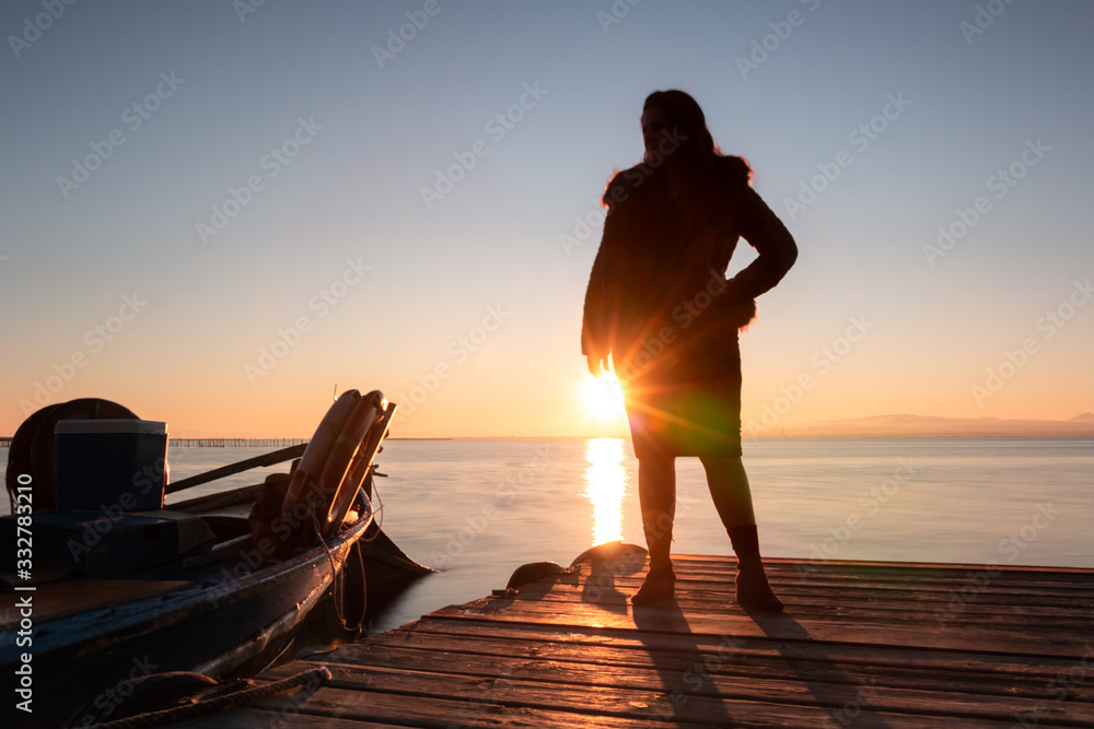 Silhouette of a woman looking at the horizon against the sunlight on a jetty of a lake at sunset