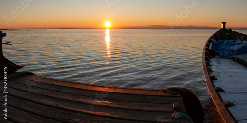Panoramic view at ground level of a wooden jetty with a wooden boat with the sun setting on the horizon in the coastal lagoon of Valencia