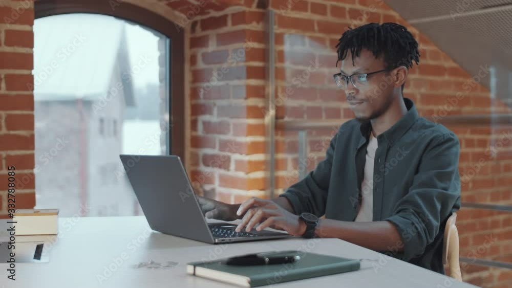 Young African American man with braid hairstyle typing on laptop while working at desk near arch top window in loft office with brick and glass walls Young African American man with braid hairstyle typing on laptop while working at desk near arch top window in loft office with brick and glass walls