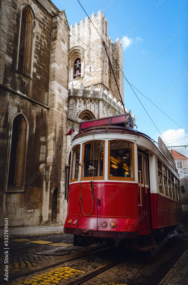 Traditional old red tramcar cable electric trolley running over the streets of Alfama historical district of Lisbon, Portugal. In background the bell tower of the nearby cathedral