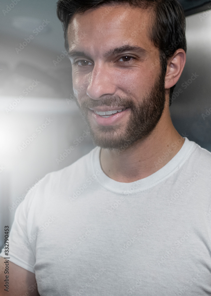 Obraz premium A close up of a tan bearded man with brown parted hair smiling through the beam of sunlight shining through the window behind him as he leans up against the refrigerator wearing a white shirt.