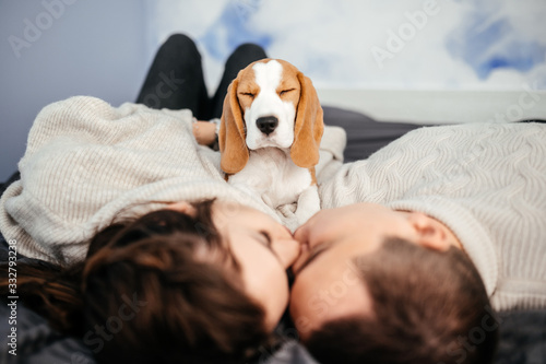 Loving couple is kisses while lying on the couch and their funny beagle sits on them with their eyes closed photo