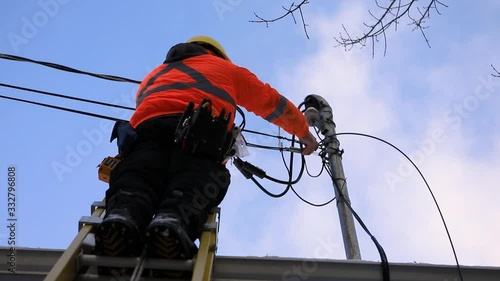 A telecoms operative man is seen working from a ladder on a utility pole, wearing high visibility personal protective clothing, high viz PPE, and hard hat