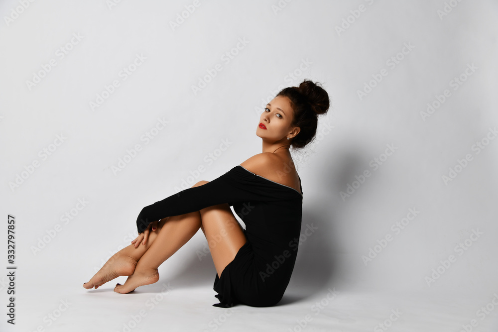 Fototapeta premium Portrait of gorgeous woman in stylish black tight dress sitting sideways to us with her legs tucked up and looking at us