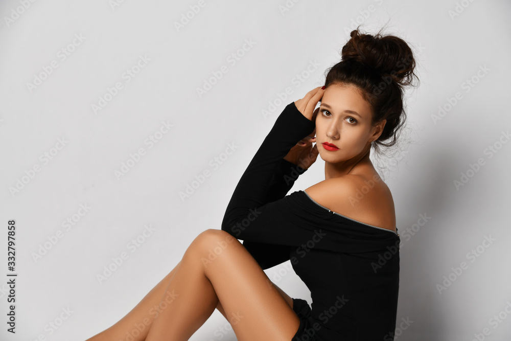 Portrait of gorgeous woman in stylish black tight dress sitting sideways to us with her legs tucked up and looking at us