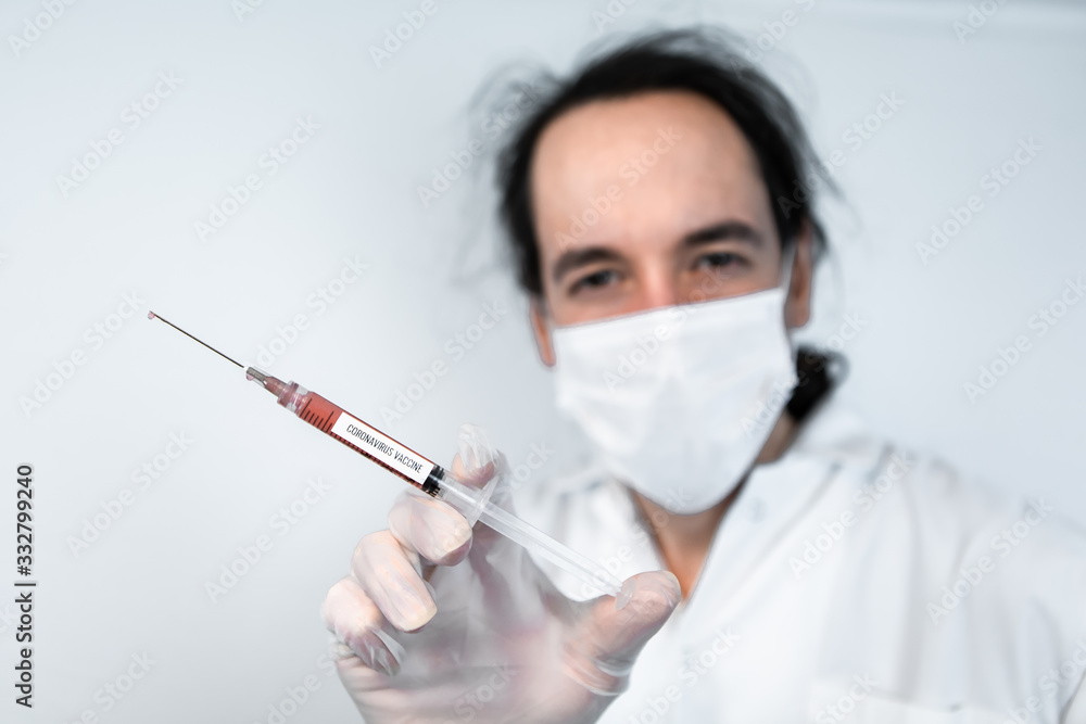 Selective focus shot of syringe with red fluid labeled coronavirus vaccine, held by gloved hand of out of focus masked scientist over a white background Selective focus shot of syringe with red fluid labeled coronavirus vaccine, held by gloved hand of out of focus masked scientist over a white background
