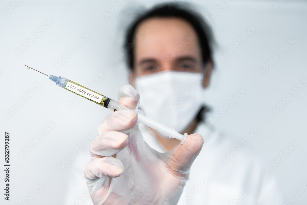 Selective focus closeup shot of syringe labeled 2019-nCOV vaccine held by gloved hand of out of focus masked scientist over a white background Selective focus closeup shot of syringe labeled 2019-nCOV vaccine held by gloved hand of out of focus masked scientist over a white background
