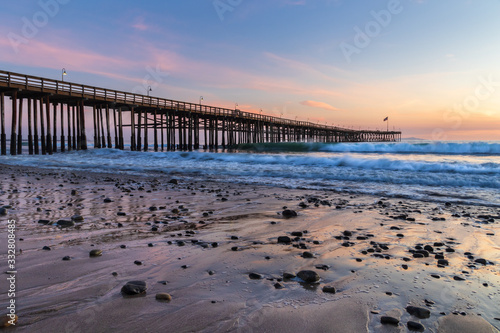 Ventura Pier at sunset  Ventura  California. Rocks and sand in foreground  water receded in low tide  with incoming waves. Lamps on pier  colored twilight sky in background.