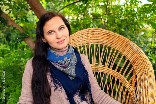 Woman in blue headscarf and in a jacket sitting on a braided chair in the garden