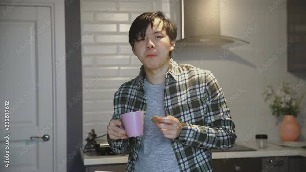 Close up of sick asian man eating cookie and drinking coffee in kitchen at home Close up of sick asian man eating cookie and drinking coffee in kitchen at home