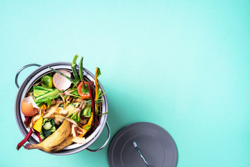 Top view of kitchen food waste collected in recycling compost pot. Peeled vegetables on chopping board, white compost bin on blue background.