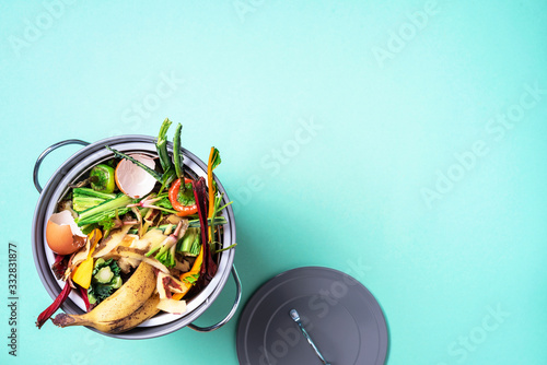 Top view of kitchen food waste collected in recycling compost pot. Peeled vegetables on chopping board, white compost bin on blue background.